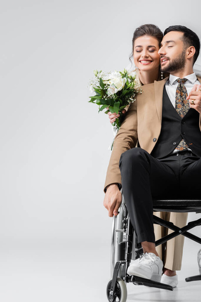 bruid met bruiloft boeket en moslim man in rolstoel hand in hand met gesloten ogen op grijs - Foto, afbeelding