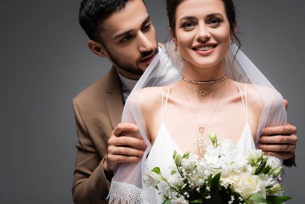 χαρούμενη, κομψή νύφη με γαμήλιο μπουκέτο, κοντά στο αραβικό γαμπρό απομονωμένο σε γκρι - Φωτογραφία, εικόνα