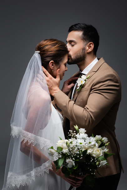 μουσουλμάνος άνδρας φιλιά νύφη κρατώντας γαμήλιο μπουκέτο απομονώνονται σε γκρι - Φωτογραφία, εικόνα