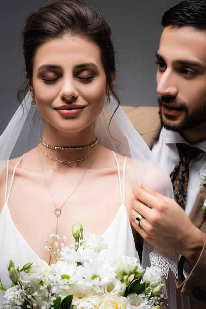 ευτυχισμένη, κομψή νύφη με κλειστά μάτια κρατώντας γαμήλιο μπουκέτο κοντά στον μουσουλμάνο γαμπρό απομονωμένο σε γκρι - Φωτογραφία, εικόνα