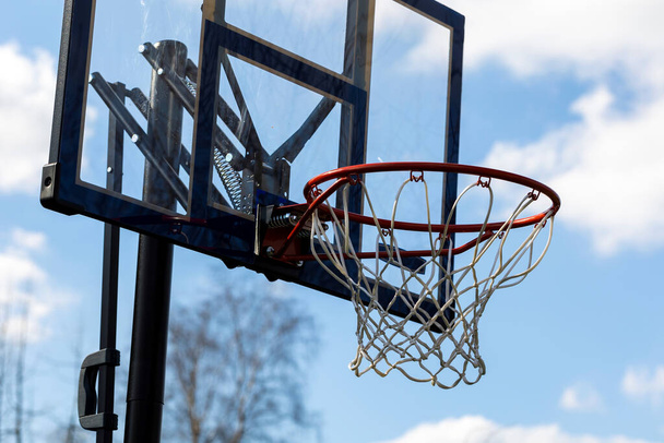 Das Porträt eines Freizeit-Outdoor-Basketballfelges mit Netz und einem Plexiglas-Backboard mit blauen Linien vor blauem Himmel. Der Basketballkorb oder Ring ist orange und seine Stange ist schwarz. - Foto, Bild