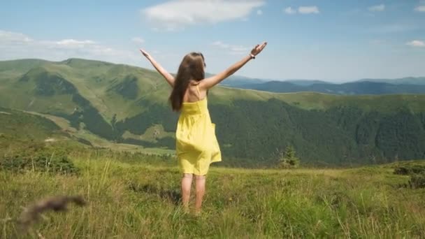 Back view of young happy woman traveler in yellow dress standing on grassy hillside on a windy day in summer mountains with outstretched arms enjoying view of nature. - Footage, Video