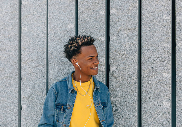 Усміхнений молодий чоловік в джинсовій куртці і навушники біля сірої стіни
 - Фото, зображення