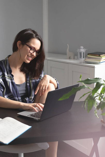 Hymyilevä nuori nainen freelancer käyttää kannettavaansa opiskeluun ja työskentelyyn verkossa kotoa nauttien mukavasta etätyöstä kotitoimistosta. Itsenäinen ammatinharjoittaminen ja freelance-käsite - Valokuva, kuva