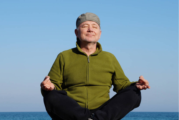 взрослый мужчина медитирует с улыбкой на фоне морского горизонта. - Фото, изображение