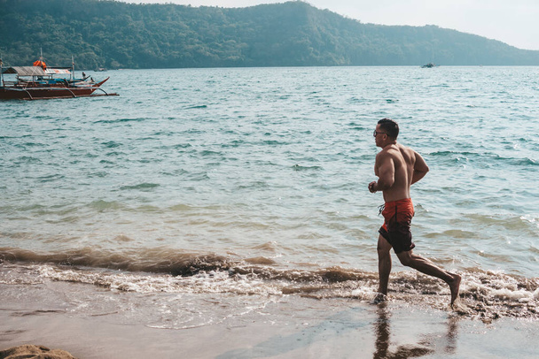 Zdolny i wysportowany mężczyzna biegnie boso wzdłuż wybrzeża. Rekreacyjne i niskiej intensywności cardio na malowniczej plaży. - Zdjęcie, obraz