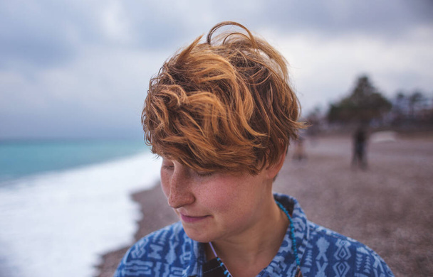 Ritratto di una ragazza dai capelli rossi con lentiggini, una donna con i capelli corti, vento tra i capelli, una passeggiata lungo la costa, una ragazza sorridente sulla spiaggia. - Foto, immagini