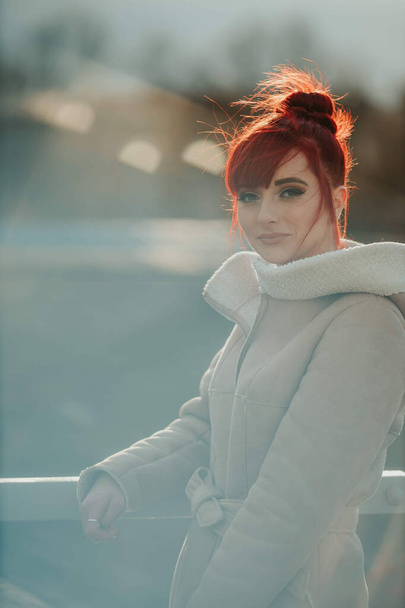 A portrait of a young smiling red-haired woman with bangs standing by the metal fence wearing a warm winter coat on a sunny day - Foto, immagini