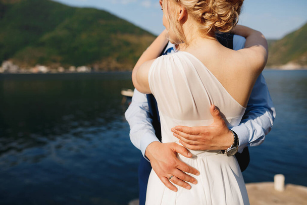 Braut und Bräutigam umarmen sich auf der Seebrücke in Perast, der Bräutigam legt die Hände auf die Taille der Braut  - Foto, Bild