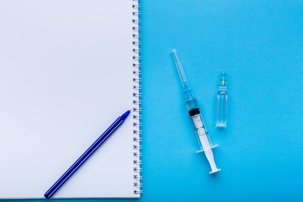 A syringe with vaccine on blue background with white blank notebook for a prescription. - Photo, Image
