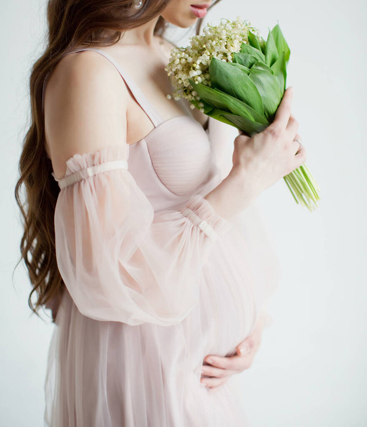 Pregnant woman in pink dress holds spring flowers lily of the valley on a white background - Photo, Image
