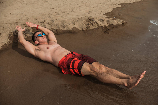 Un atlético y deportivo asiático hace V-ups en la arena en la playa. Entrenamiento abdominal o calistenia al aire libre. - Foto, imagen