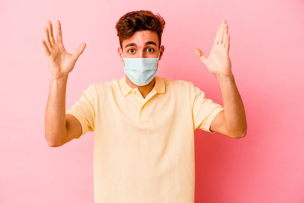 Joven hombre caucásico llevando una protección para coronavirus aislado sobre fondo rosa recibiendo una agradable sorpresa, excitado y levantando las manos. - Foto, Imagen