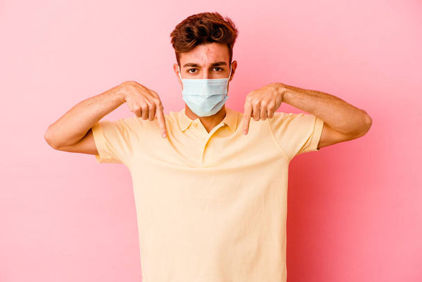 Junger kaukasischer Mann mit einem Schutz gegen Coronavirus isoliert auf rosa Hintergrund zeigt mit den Fingern nach unten, positives Gefühl. - Foto, Bild