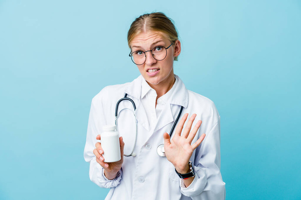 Jeune femme médecin russe tenant bouteille de pilules sur bleu rejetant quelqu'un montrant un geste de dégoût. - Photo, image