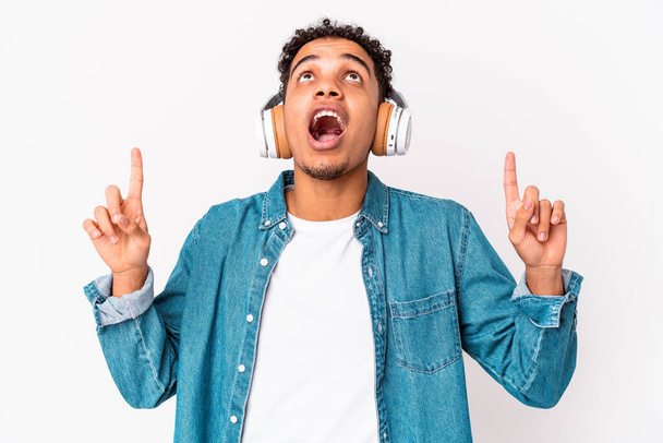 Młody, afrykański kręcony mężczyzna odizolowany słuchając muzyki ze słuchawkami skierowanymi do góry nogami z otwartymi ustami. - Zdjęcie, obraz