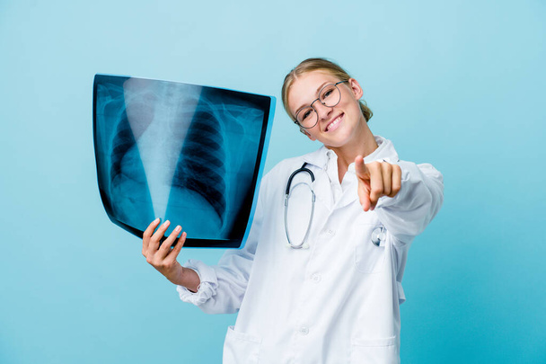 Joven doctora rusa sosteniendo un escaneo óseo en azul sonrisas alegres apuntando hacia el frente. - Foto, Imagen
