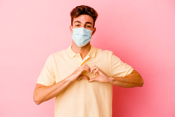 Молодий білий чоловік в захисті від коронавірусу ізольований на рожевому фоні посміхається і показує форму серця руками
. - Фото, зображення