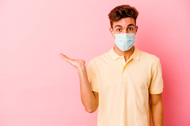 Junger kaukasischer Mann mit Coronavirus-Schutz isoliert auf rosa Hintergrund beeindruckt mit Kopierraum auf Handfläche. - Foto, Bild