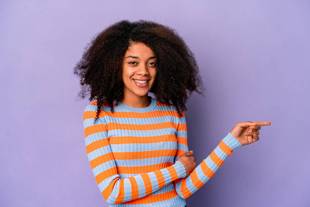 Joven africana americana rizada mujer aislada sobre fondo púrpura sonriendo alegremente señalando con el dedo índice de distancia. - Foto, Imagen