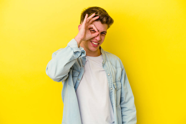 Νεαρός καυκάσιος άνδρας απομονωμένος σε κίτρινο φόντο ενθουσιασμένος κρατώντας εντάξει χειρονομία στο μάτι. - Φωτογραφία, εικόνα