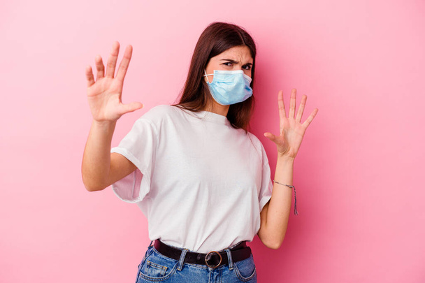 Mujer caucásica joven que lleva una máscara para el virus aislado en el fondo rosa se sorprende debido a un peligro inminente - Foto, Imagen