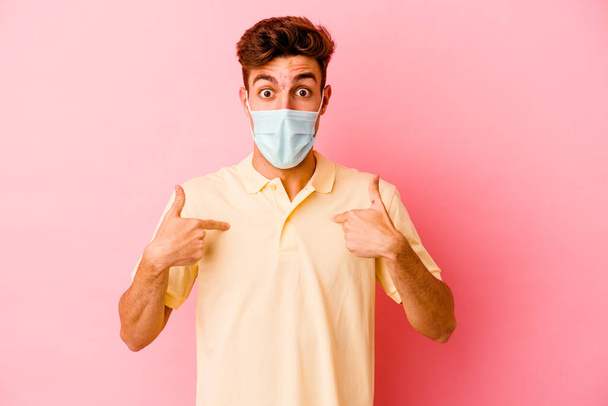 Jeune homme caucasien portant une protection contre le coronavirus isolé sur fond rose surpris pointant du doigt, souriant largement. - Photo, image
