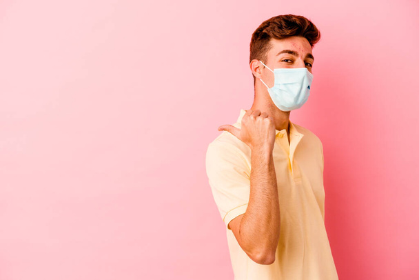 Fiatal fehér férfi visel védelmet a koronavírus izolált rózsaszín háttér pontok hüvelykujj el, nevetve és gondtalanul. - Fotó, kép