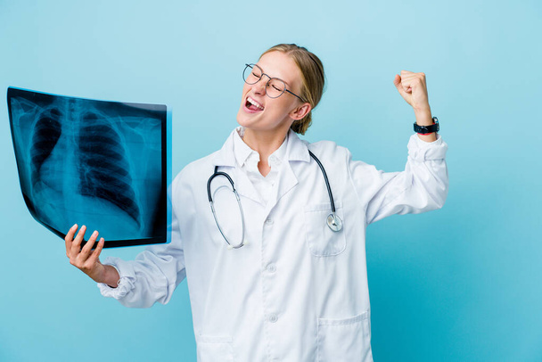 Young russian doctor woman holding a bone scan on blue raising fist after a victory, winner concept. - Photo, Image
