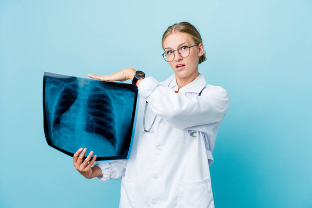 Young russian doctor woman holding a bone scan on blue shocked and amazed holding a copy space between hands. - Photo, Image