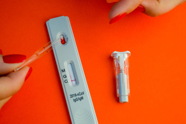 test op coronavirus op een oranje achtergrond. op een matte achtergrond een gevoelige strip, een meisje met een felrode manicure druipt op een druppel bloed voor een test. - Foto, afbeelding