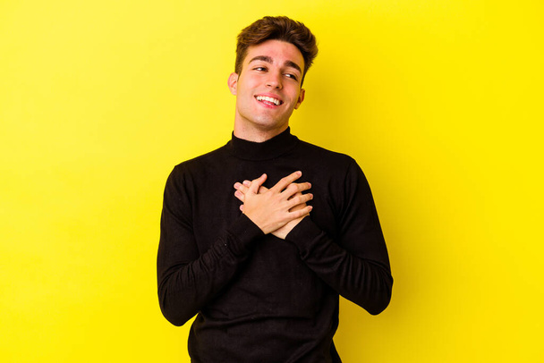 Молодой кавказский человек изолирован на желтом фоне смеясь держа руки на сердце, понятие счастья. - Фото, изображение