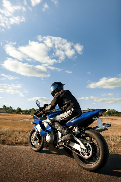 Motosikletli ya da motosikletli siyah deri takım elbiseli ve kasklı bir sporcu motosikletine biner ve uzaklığa bakar. Dikey fotoğraf yönelimi - Fotoğraf, Görsel