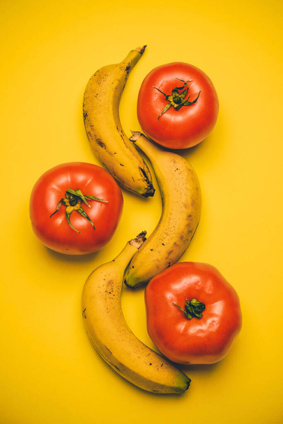 黄色の背景に熟した赤いトマトが散在している3つの黄色の熟したバナナが並んでいます。ビーガンコンセプト。果物だ。健康食品. - 写真・画像
