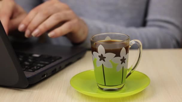 Una taza de café en primer plano. La joven trabaja detrás de un portátil en el fondo y periódicamente recoge una taza de bebida con su mano. - Metraje, vídeo