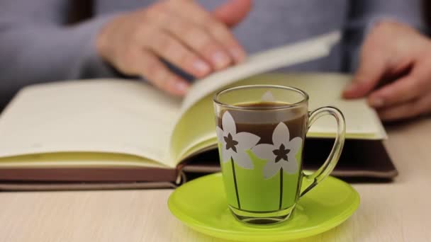 Cup of coffee in the foreground. The young woman leafing through a book, notebook in the background and periodically picks up a cup of drink with his hand. - Footage, Video