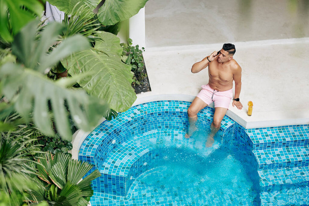 Serious fit man sunbathing by swimming pool, drinking orange juice and talking on phone - 写真・画像