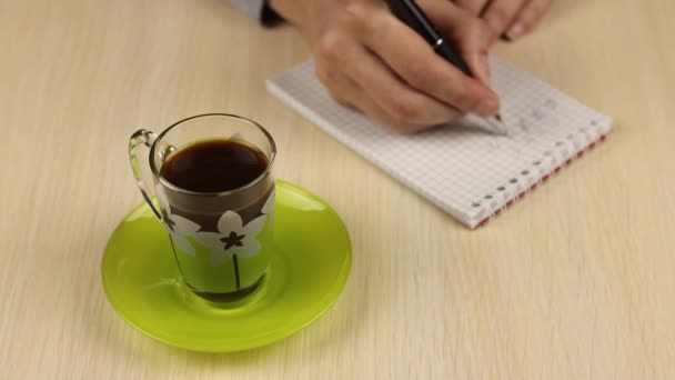 Ένα φλιτζάνι καφέ σε πρώτο πλάνο. Η νεαρή γυναίκα γράφει εργασίες σε ένα σημειωματάριο στο παρασκήνιο. και παίρνει περιοδικά ένα φλιτζάνι ποτό με το χέρι του. - Πλάνα, βίντεο