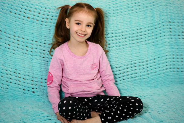 Kis boldog lány aranyos copfokkal ül egy kék kanapén és mosolyog. A gyermek örül, és jó szórakozást pózol a fotósnak. Szabad hely a szövegnek. Egy boldog gyermek portréja - Fotó, kép