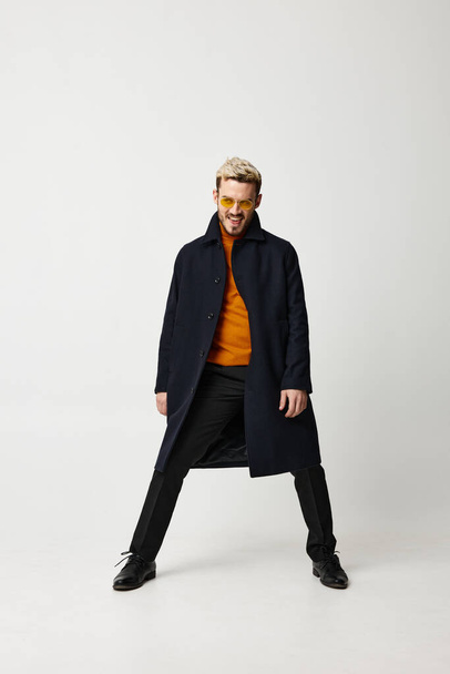 модный мужчина в пальто раздвинул ноги на светлом фоне и оранжевый свитер брюки туфли - Фото, изображение