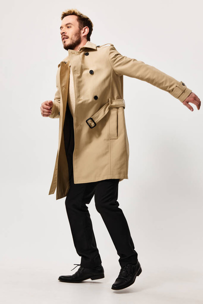a guy in a beige coat gestures with his hands on a light background in full growth side view - Photo, Image