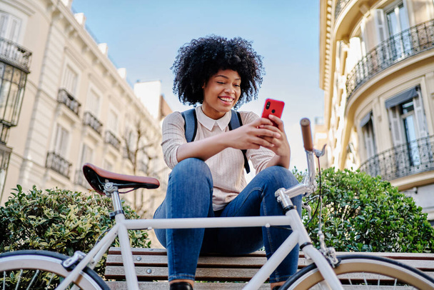 Afro saçlı Latin bir kadın bankta oturmuş cep telefonuna bakıyor. - Fotoğraf, Görsel