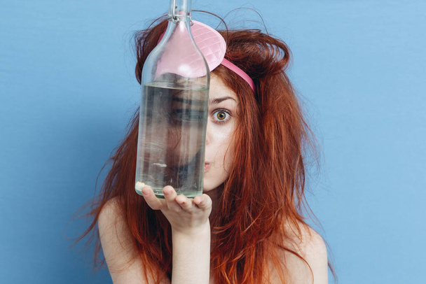 woman with tousled hair holding bottle of alcohol near face blue background - Foto, afbeelding