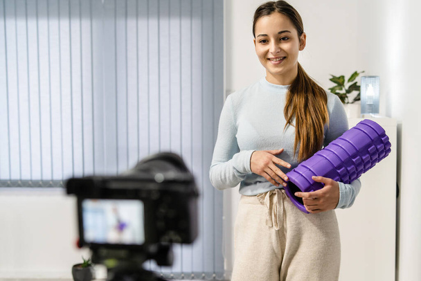 Вид спереди на молодую кавказскую женщину дома, запись и потоковое онлайн видео на камеру для онлайн-тренировок - Девушка, использующая пенопласт для тренировок в комнате - концепция здоровья и фитнеса - Фото, изображение
