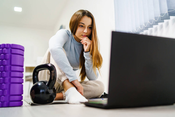 トレーニングのために自宅でオンラインビデオを見て床に座っている若い白人女性のフロントビュー-インターネット上でワークアウトを探しているラップトップコンピュータを持つ女の子-健康とフィットネスのコンセプトコピースペース - 写真・画像