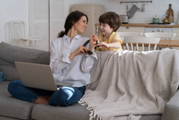 Madre freelancer sentada en el sofá en casa, trabajando en el ordenador portátil, niño distraído y pidiendo atención - Foto, imagen