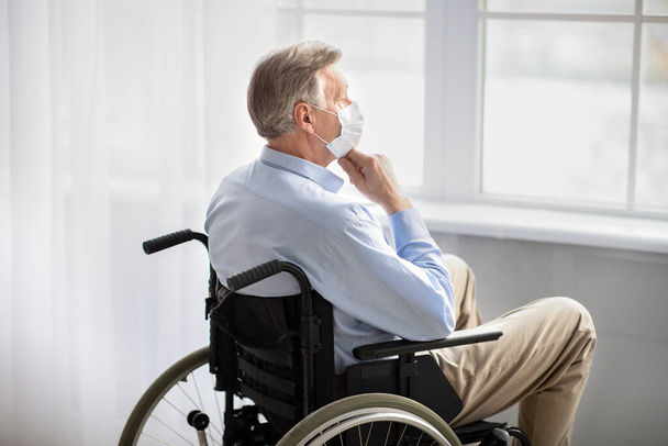 Zdravotně postižení senioři během covidské karantény. Osobně postižený starší muž v masce se dívá z okna domů, cítí depresi - Fotografie, Obrázek