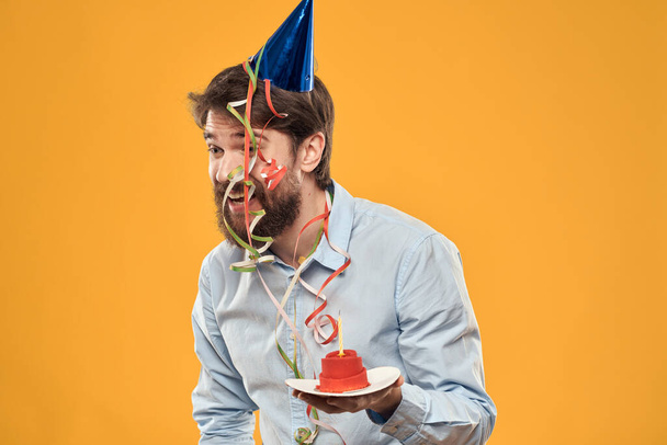 Homme joyeux avec un gâteau sur un fond jaune casquette de vacances d'anniversaire sur sa tête - Photo, image
