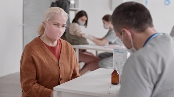 Średni POV blondynki starszej rasy białej kobiety w różowej masce na twarzy, siedzącej przy biurku u lekarza, który pracuje. Skrzyżowanie ramion pacjenta na klatce piersiowej i patrzenie na kamerę - Materiał filmowy, wideo