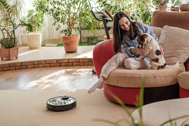 Роботизированный пылесос на ковре, женщина в голубой рубашке играет с собакой, Джек Рассел терьер породы дома на диване,, наслаждаясь жизнью концепции, уютный и уютный дом, смарт-устройство - Фото, изображение